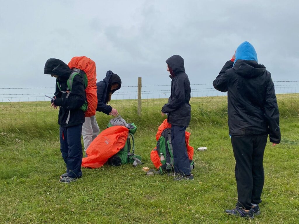 Students with hoods up putting on the waterproof material for their rucksacks