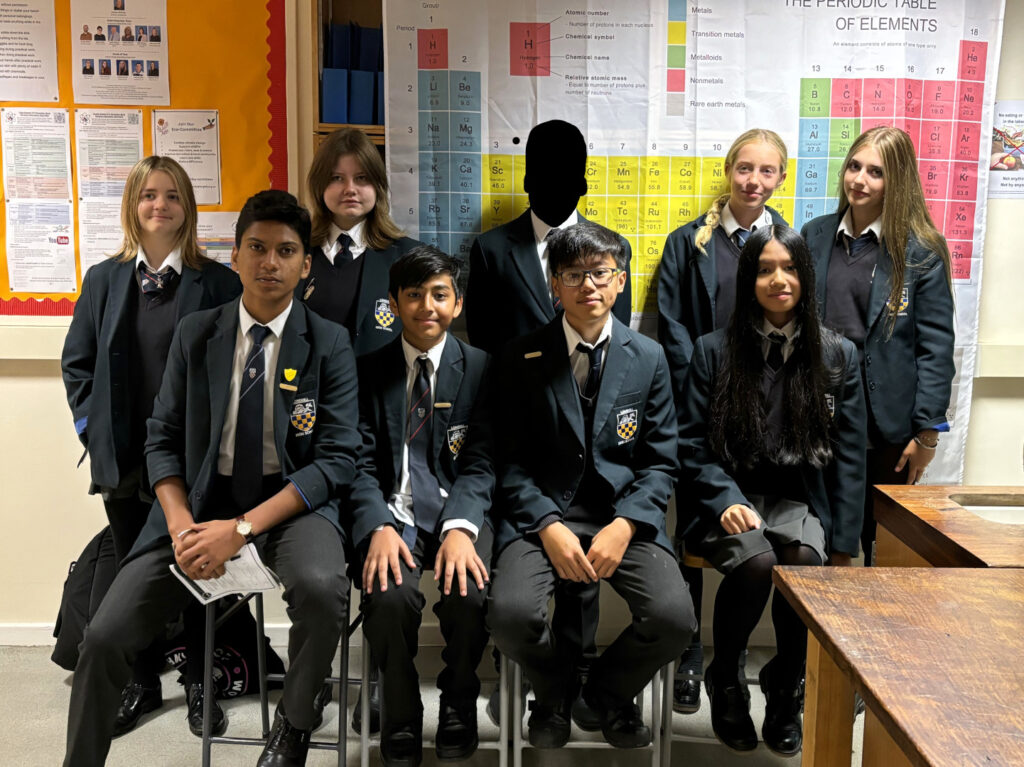 A group of students posing for a picture in a science classroom. The periodic table of elements is on a poster behind them. 9 students are gathered in the picture.