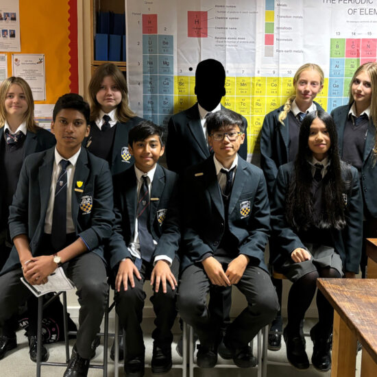 A group of students posing for a picture in a science classroom. The periodic table of elements is on a poster behind them. 9 students are gathered in the picture.